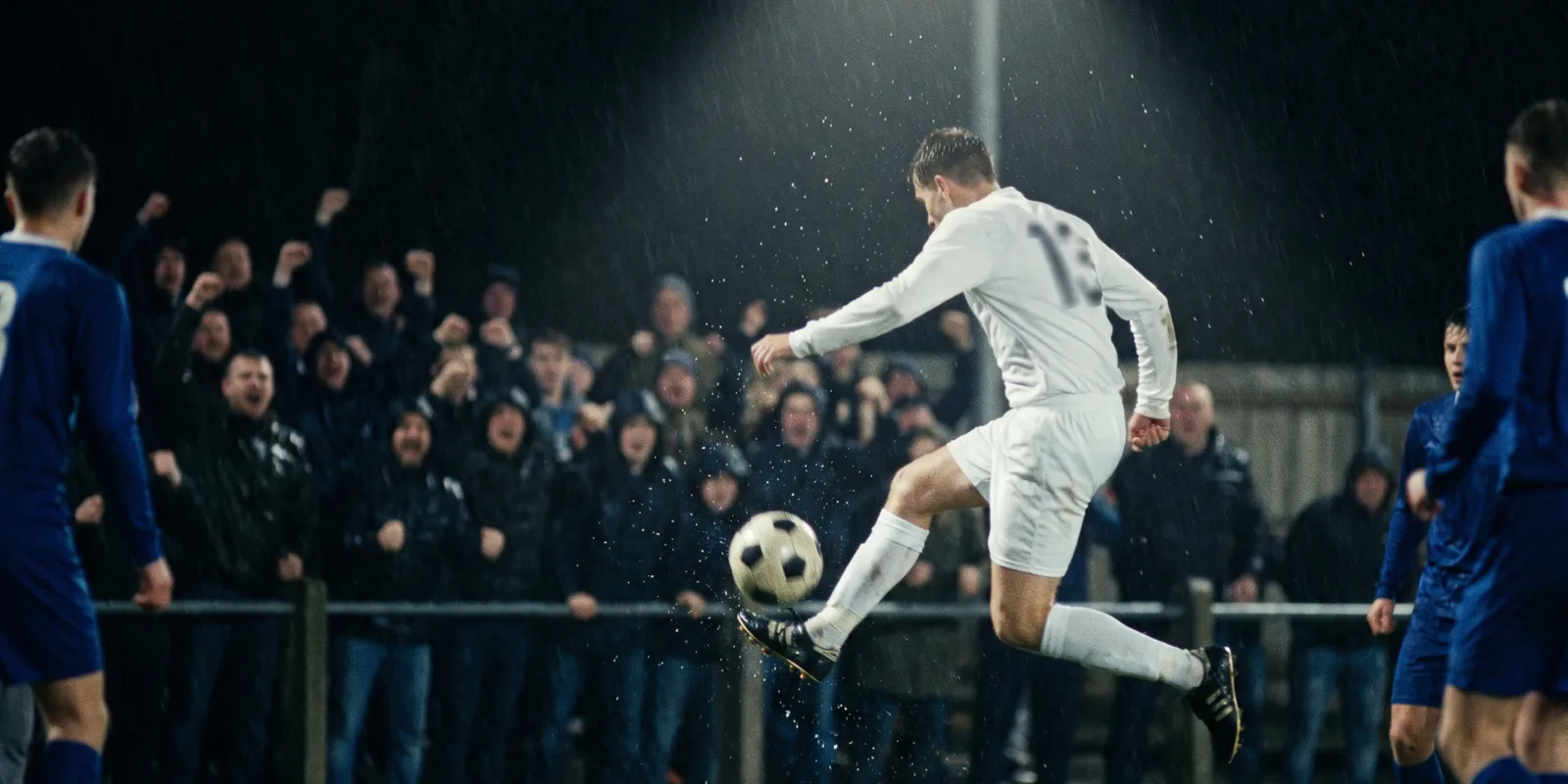 A young footballer striking the ball under floodlights, rain sparkling in the beams.